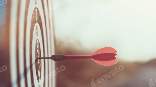 Preview: Close up red dart arrow hitting target center dartboard on sunset background.