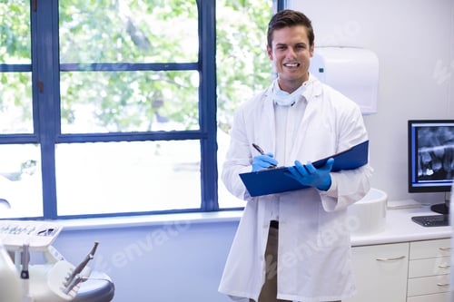Preview: Male dentist standing by window holding clipboard near dental chair in dental clinic, copy space