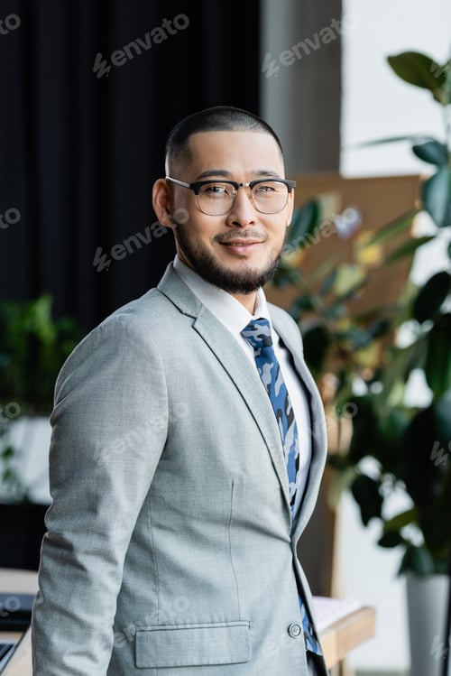 Preview: happy asian businessman in formal wear and eyeglasses looking at camera in office