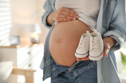 Preview: Young pregnant woman with baby's shoes indoors, closeup. Space for text