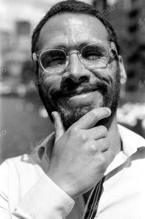Preview: Smiling bearded man wearing glasses is touching his chin in black and white analog photo