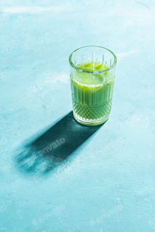 Preview: Vertical shot of a transparent glass containing a bright green healthy liquid on a blue surface