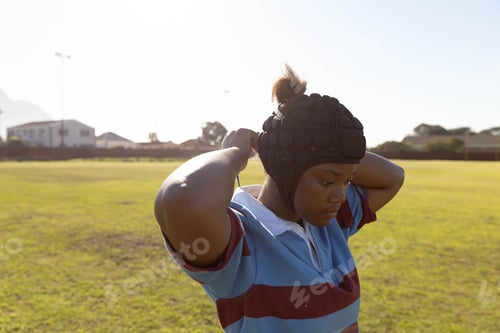 Preview: African American teenage girl wearing rugby jersey adjusting padded headguard on grass field