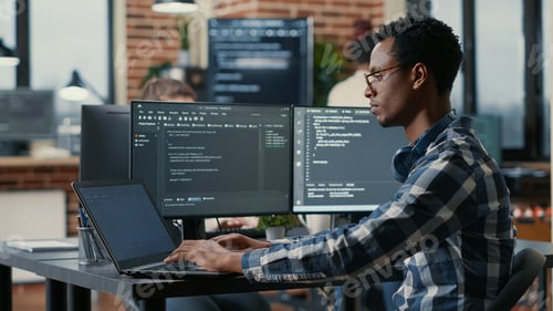 Preview: Portrait of african american developer using laptop to write code sitting at desk with multiple
