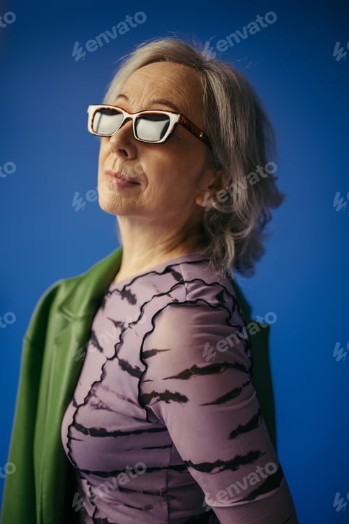 Preview: portrait of senior woman in sunglasses standing with green leather jacket isolated on blue