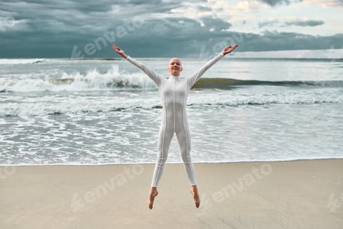 Preview: Joyful hairless girl with alopecia in white futuristic suit jumping sea sandy beach