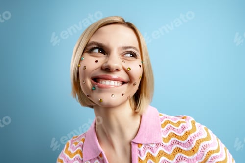 Preview: Playful young woman with colorful rhinestones over her face smiling against blue background
