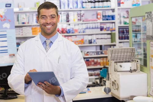 Preview: Male pharmacist in lab coat holding and using tablet behind pharmacy counter with pill dispenser