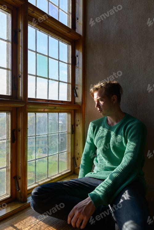 Preview: Young handsome Caucasian man sitting in front of closed wooden window with sunlight streaming in