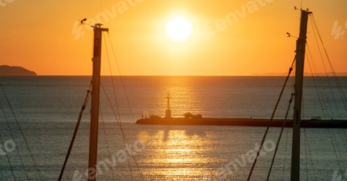 Preview: Sunset over Tinos island, Cyclades, Greece. View of beacon silhouette between ship mast, golden sun.