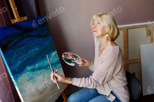 Preview: Happy blonde mature female painting on canvas at home. Looking at camera.