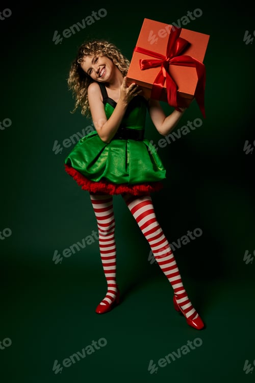 Preview: beautiful curly woman in green dress smiling cheerfully and raising huge gift, new year elf concept