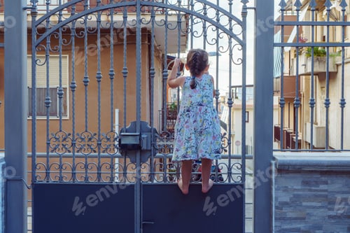 Preview: Back view tanned barefoot little girl wearing blue summer dress climbing wrought iron closed gates
