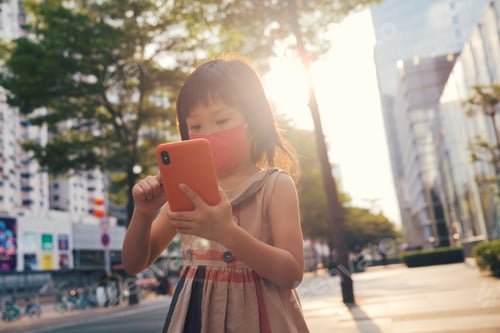 Preview: Asian child wearing mask and using cell phone in city