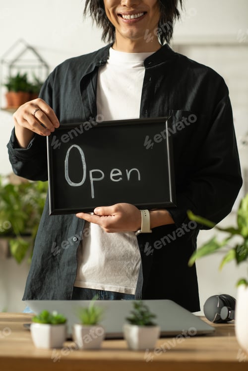 Preview: Young asian florist welcoming customers with a cheerful smile and open sign in a vibrant shop