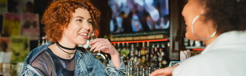 Preview: Cheerful redhead woman talking to blurred african american friend in bar, banner