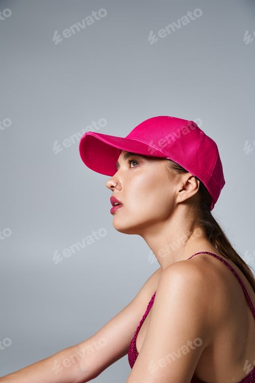 Preview: Side view of beautiful brunette woman in trendy attire and pink cap in studio setting