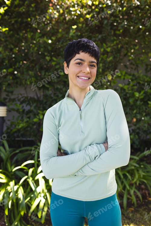 Preview: Portrait of happy biracial woman with short dark hair crossing arms and smiling in sunny garden