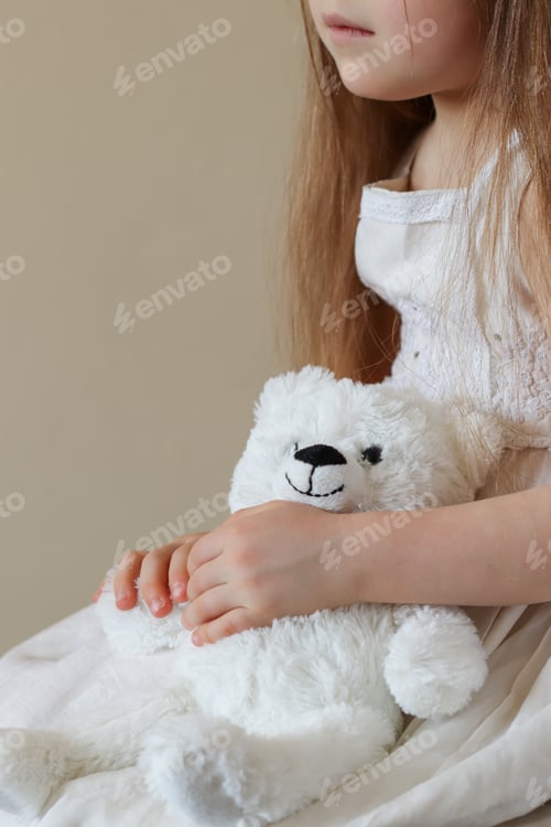 Preview: A little girl in a white dress holds a teddy bear in her hands
