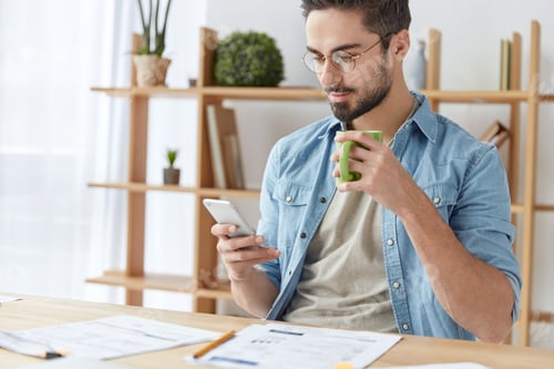 Preview: Busy young hardworking male sits at working desk, holds smart phone, view photos on Internet, drinks