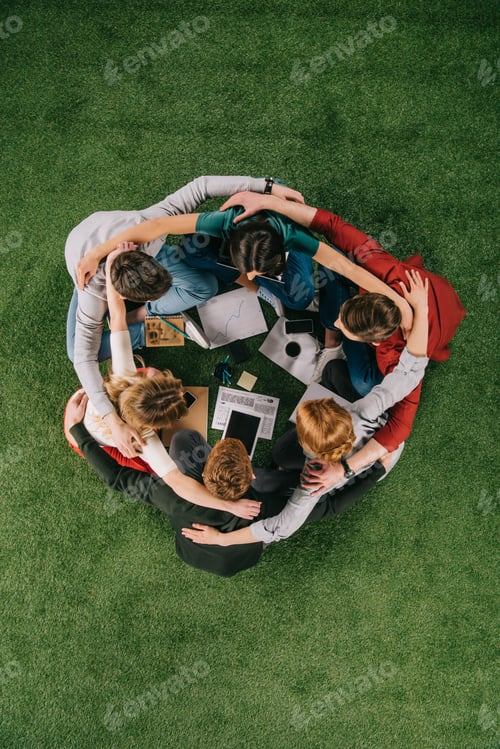 Preview: overhead view of businesspeople embracing while sitting on grass with devices and documents