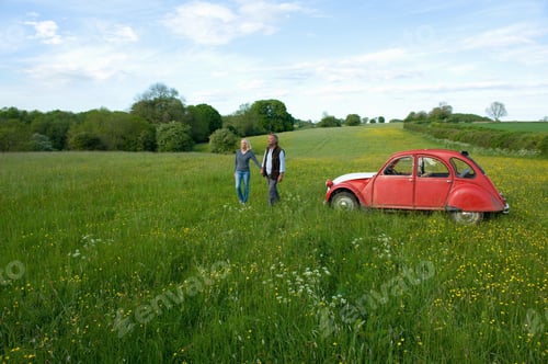 Preview: Man and woman walking hand in hand across a meadow, vintage red car parked close by.