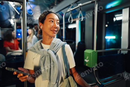 Vista previa: Young woman using public transport at night holding smartphone and wearing wireless earbuds