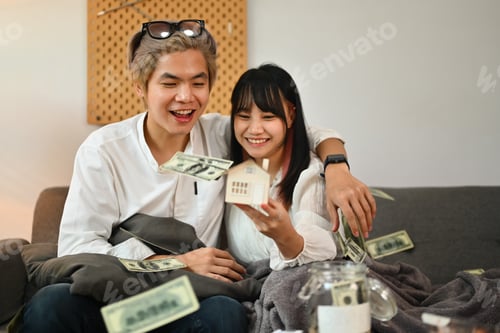 Preview: Excited young couple holding house model sitting on couch with dollar banknotes falling from above