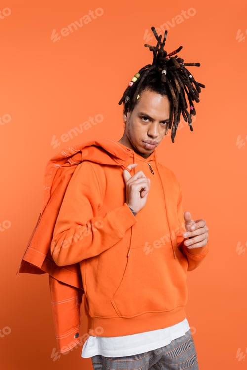 Preview: tattooed and multiracial man in hoodie holding denim jacket isolated on coral background