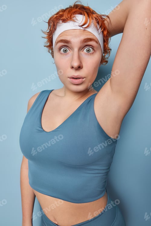 Preview: Shocked sporty woman with red hair stares impressed at camera keeps arm raised stands sweaty after