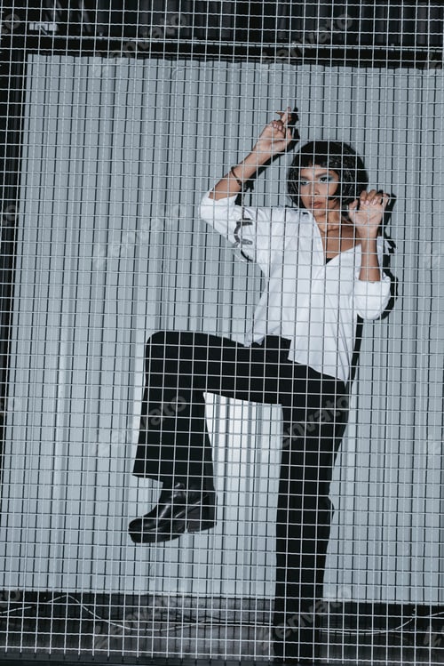 Preview: woman in white shirt with cuffs standing behind grate and looking away