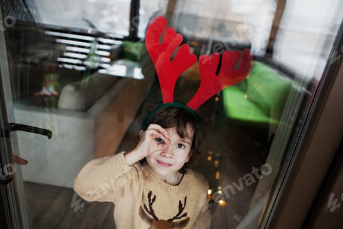 Preview: Adorable little boy wearing reindeer antlers at home and looking at camera against background of