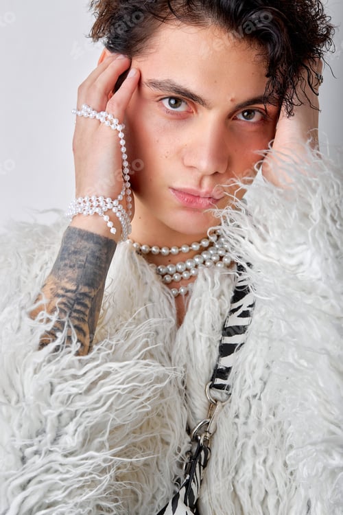 Preview: brunette man in fluffy coat and necklace looking at camera, touching temples