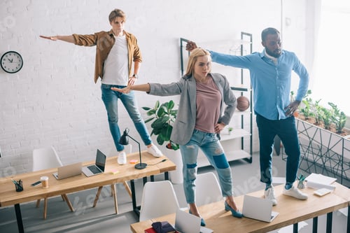 Preview: high angle view of happy multiethnic business colleagues having fun and dancing on tables in modern