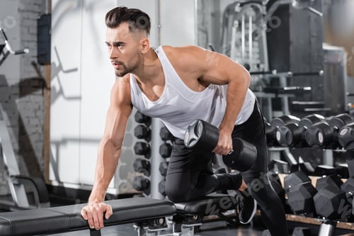 Preview: Young sportsman working out with dumbbell on flat bench in gym