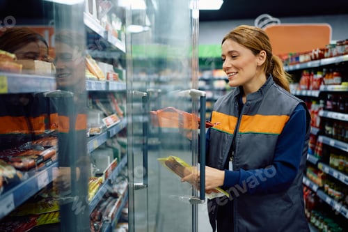 Preview: Happy supermarket worker stacking products in a refrigerator at the store.