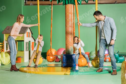 Preview: smiling parents looking at cute happy children swinging on swings in game center