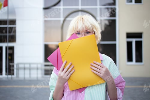 Preview: Funny teenage girl outside holding student hides face behind folders.
