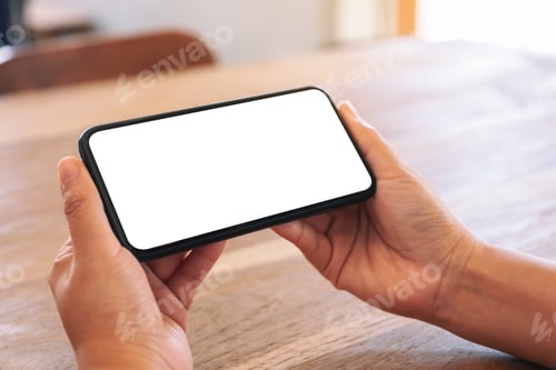 Preview: Hands holding mobile phone with blank white screen horizontally on wooden table