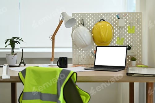 Preview: Architect working desk with laptop computer, hardhat, reflective jacket and stationery.