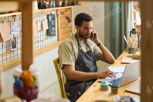 Preview: Young male clerk in apron calling clients and looking through online orders