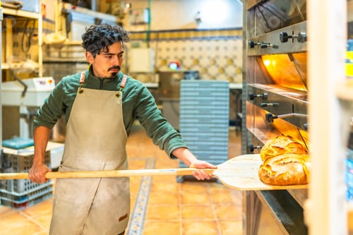 Preview: Man using peel to remove baking bread from traditional oven