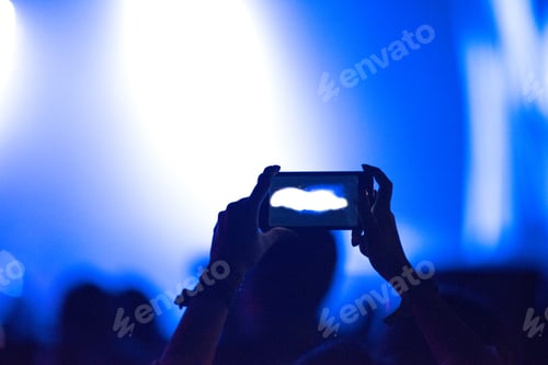Preview: Hand holding smart phone at live music festival. Concert stage lights on viewfinder