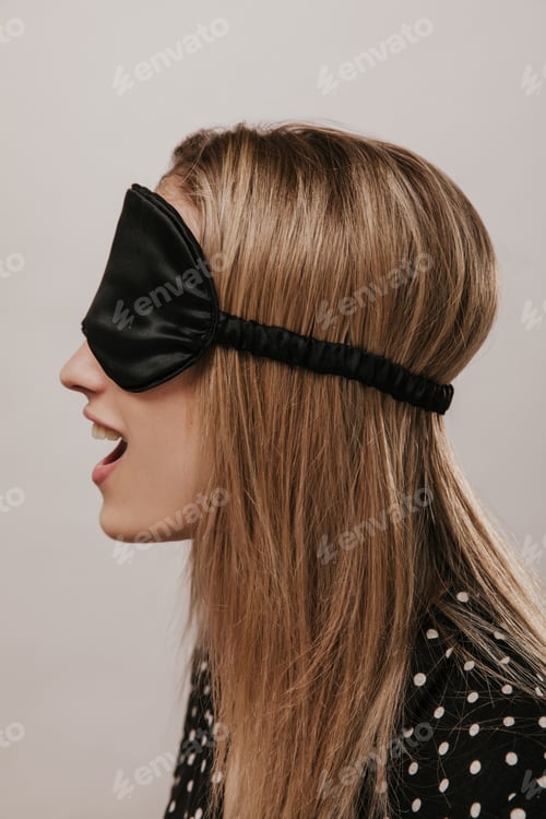 Preview: Excited young lady with blonde hair, black polka dot shirt and silk sleeping mask, opening mouth an