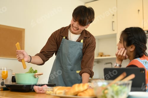 Preview: Shot of young man putting raw spaghetti pasta into pot while cooking with his wife in kitchen