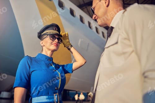 Preview: Charming woman flight attendant in hat and suit welcoming pilot near the airplane