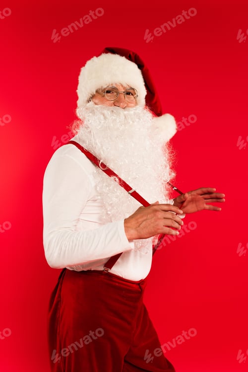 Preview: Santa claus in hat and eyeglasses holding suspenders isolated on red