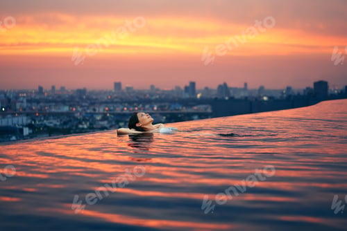 Preview: Young woman relax in swimming pool on roof top during amazing sunset and enjoy cityscape