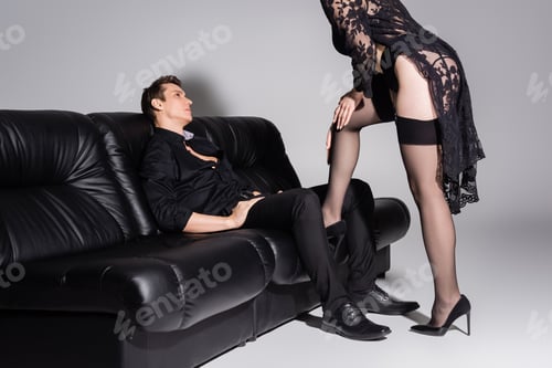 Preview: man in formal wear sitting on couch near passionate woman in black stockings on grey background