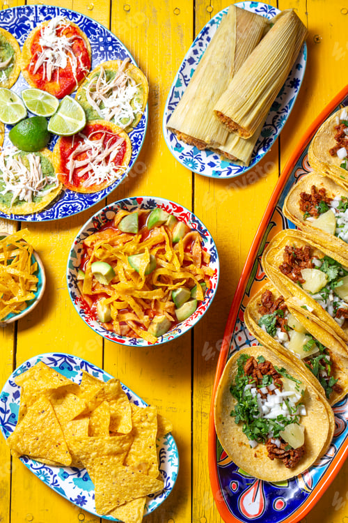 Preview: Assortment of colorful Mexican dishes on a vibrant yellow table, featuring tacos, tamales, aztek